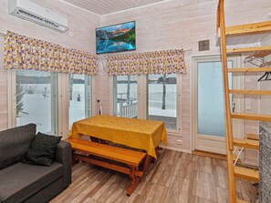 A room with a wooden table, a sofa, a TV mounted on the wall, and a window with a view of mountains.