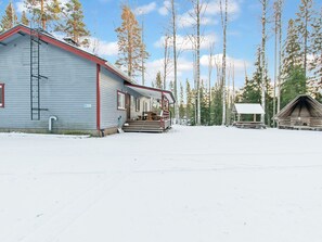 A cabin with a porch and a small attached structure, surrounded by snow and tall trees.
