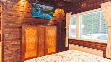 A room with wooden walls, a bed with a floral bedspread, a mounted TV, and a window with curtains.