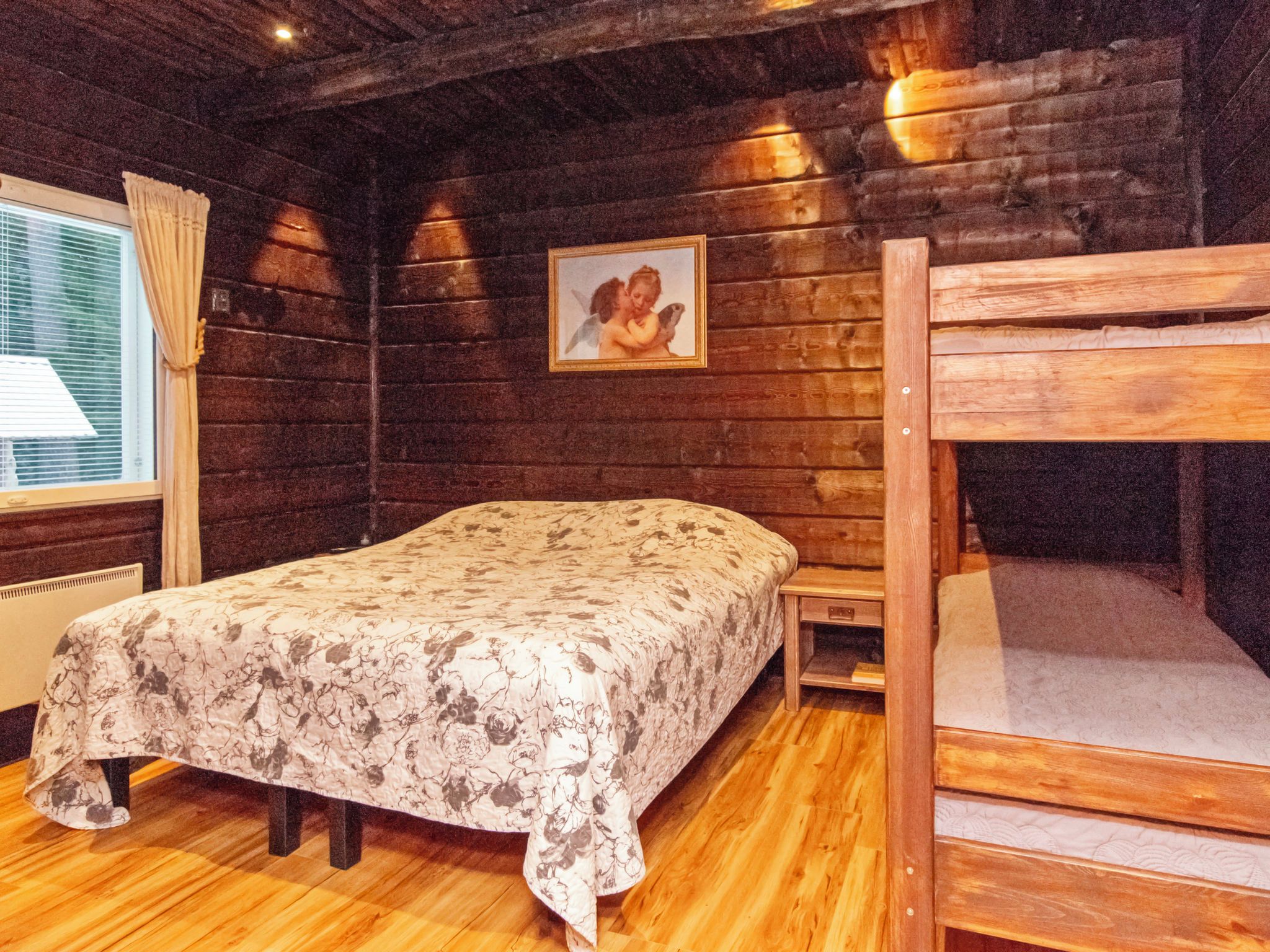 A wooden cabin room with a bunk bed, a single bed with a floral bedspread, a window with curtains, and a framed picture on the wall.