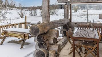 Una terrassa de fusta a l'exterior amb una barbacoa i zona d'estar, envoltada per terra i arbres coberts de neu.
