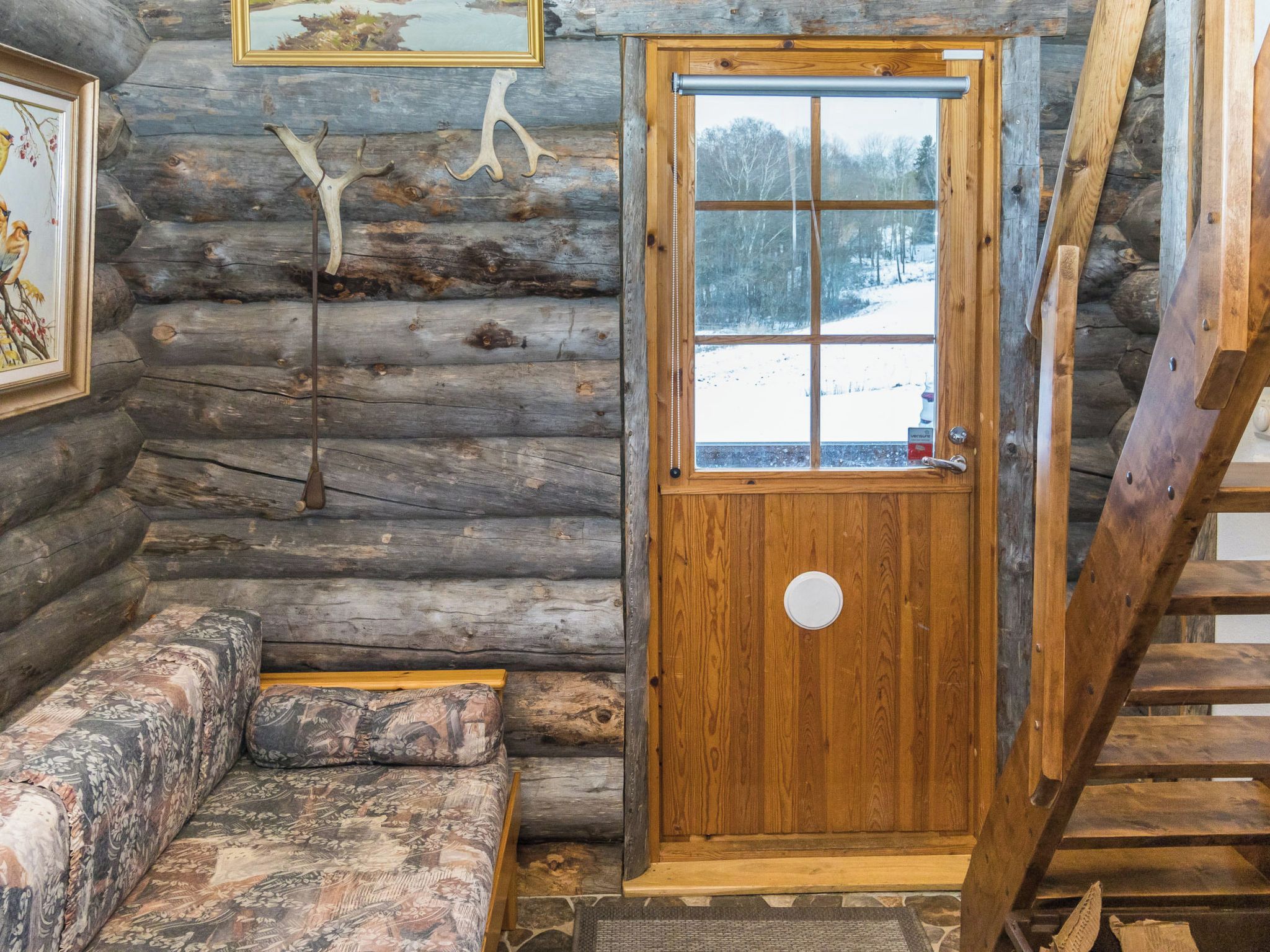 A log cabin interior with a wooden door, a bench, and a window overlooking a snowy landscape.