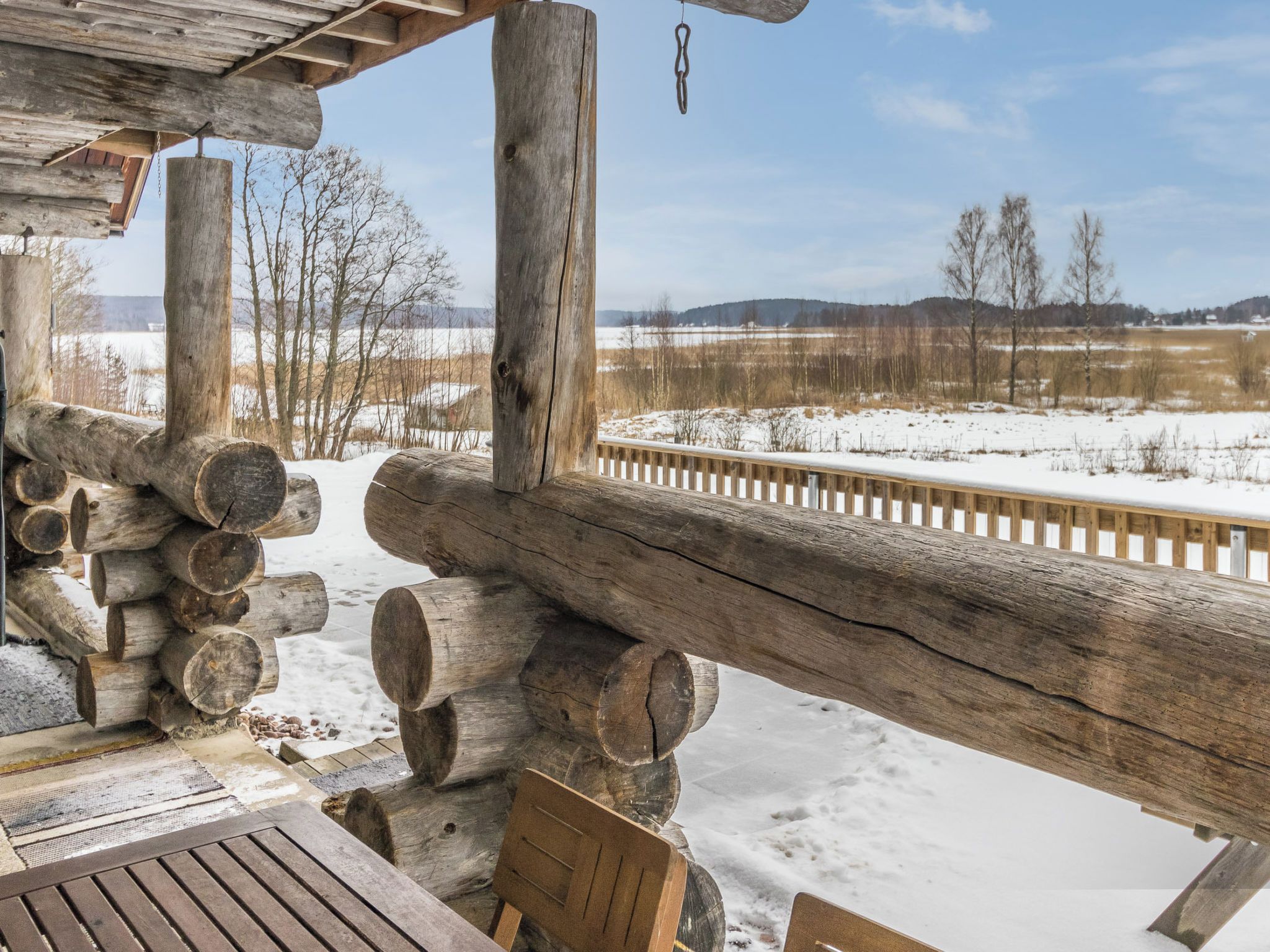 Een rustiek houten terras met een bankje en boomstammen, met uitzicht op een besneeerd landschap met een hek en bomen.