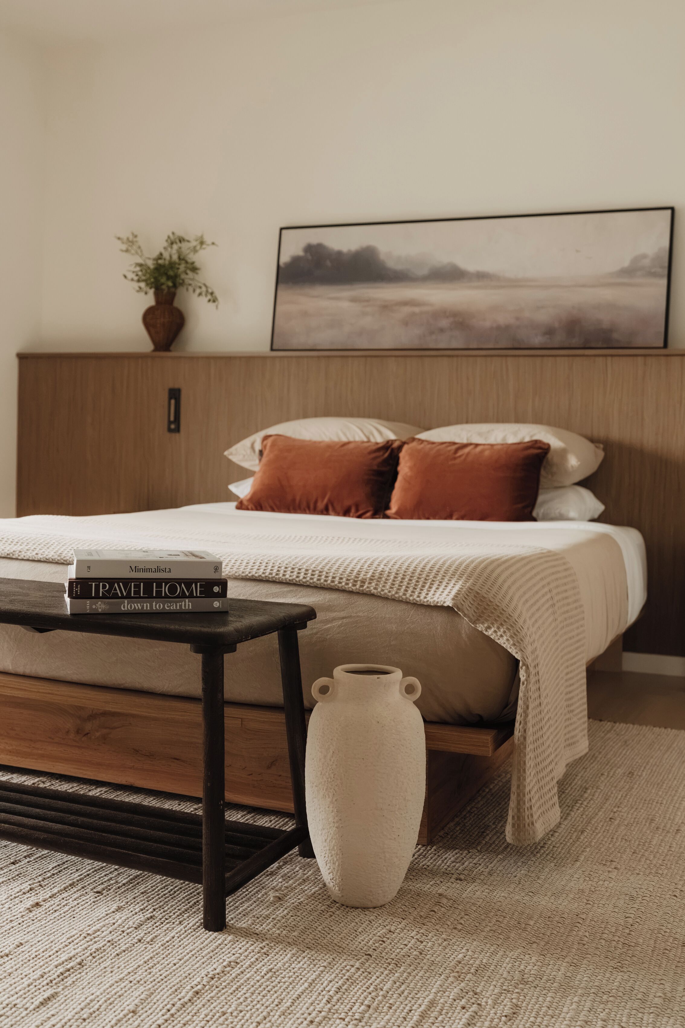A bedroom with a bed, a bedside table with books, a vase, and a painting on the wall.