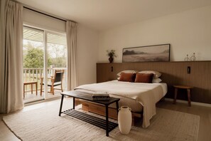 Un dormitorio con cama, mesitas de noche, un banco, un jarrón, un cuadro y vistas al exterior.