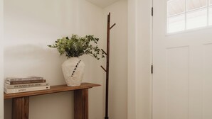 Et hjørneværelse med trægulv, en hvid dør, et knagered, en hvid vase med grønne planter og en stak bøger på et træbord.