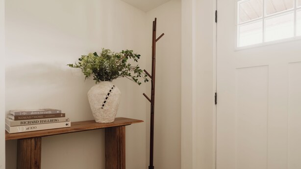 Una habitación de esquina con piso de madera, una puerta blanca, un perchero, un jarrón blanco con plantas verdes y una pila de libros sobre una mesa de madera.