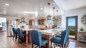 Une cuisine moderne dotée d’un grand îlot central, de tabourets de bar, d’appareils électroménagers en acier inoxydable et d’une vue sur l’extérieur, visible à travers la porte.