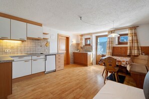 A compact kitchen with wooden cabinets, a dining area with a table and chairs, and a view through a window. - Apartment 'Wegscheiderhof' with Mountain View, Private Terrace and Wi-Fi (Dobbiaco)