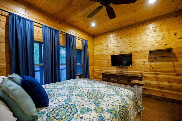 Une chambre dans un chalet en bois, avec un lit, des rideaux, un ventilateur de plafond, une télévision et une fenêtre.