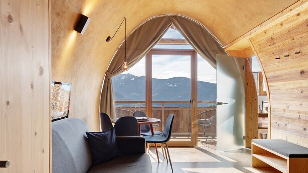 A cozy cabin interior with a wooden interior, a sofa, a dining table, and a view of mountains through a large window. - Studio 'Pavilion Water' with Mountain View, Shared Terrace and Wi-Fi (Velturno)