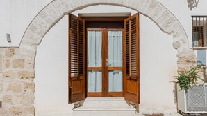 Une porte en arc surmontée de volets en bois, un mur blanc et une petite marche menant à l’entrée.