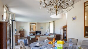 Un comedor con una araña, una mesa dispuesta con platos, tazas y una botella, y un sofá al fondo.