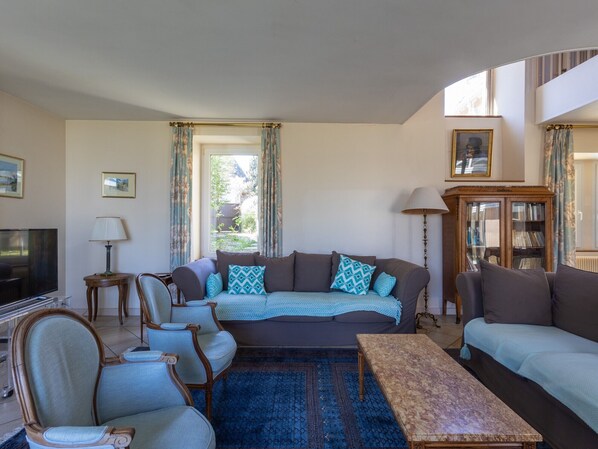 A living room with a sofa, armchairs, a coffee table, a television, a lamp, a cabinet, and a window with curtains.