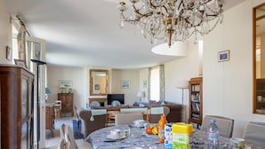 Un comedor con una araña, una mesa dispuesta con platos, tazas y una botella, y un sofá al fondo.