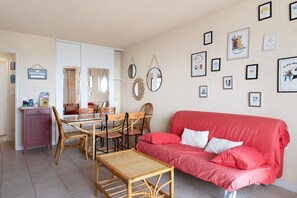A furnished living room with a red sofa, a dining table with chairs, and a kitchen area with a fridge and a cabinet.