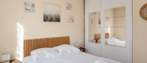 A hotel room with a bed, a bedside table, a lamp, a mirror, and wall decorations including a cityscape photo and a map.