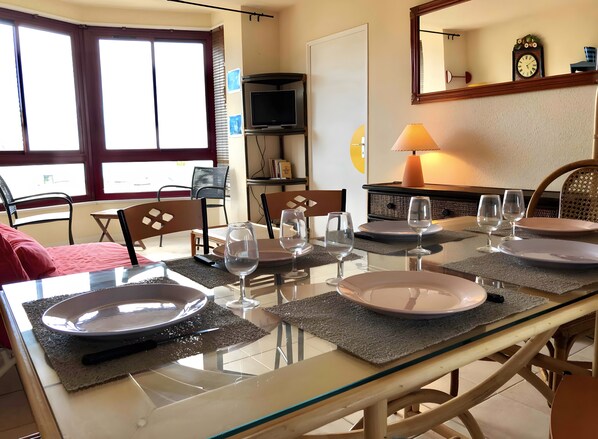 Un coin repas avec une table dressée pour quatre personnes, des chaises, un téléviseur et une horloge au mur.