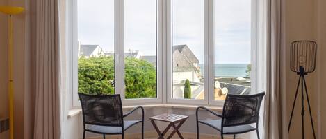 Et værelse med karnapvindue med udsigt over et boligområde og havet. Der er to sorte stole og et lille træbord.