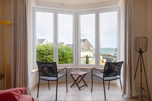 Une pièce dotée d’une grande baie vitrée donnant sur un quartier résidentiel et la mer. On y trouve deux chaises noires et une petite table en bois.