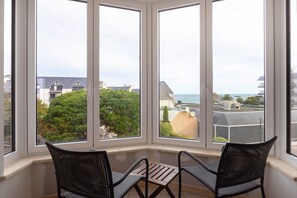 Un ventanal con dos sillones y una mesa, con vista a una zona residencial con casas y al mar.