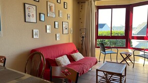 Ein Wohnzimmer mit einem roten Sofa, einem Esstisch, Stühlen, einer Stehlampe und einem großen Fenster mit Blick auf Gebäude und Grünflächen.