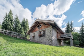 A stone house with a wooden balcony, surrounded by greenery and trees.