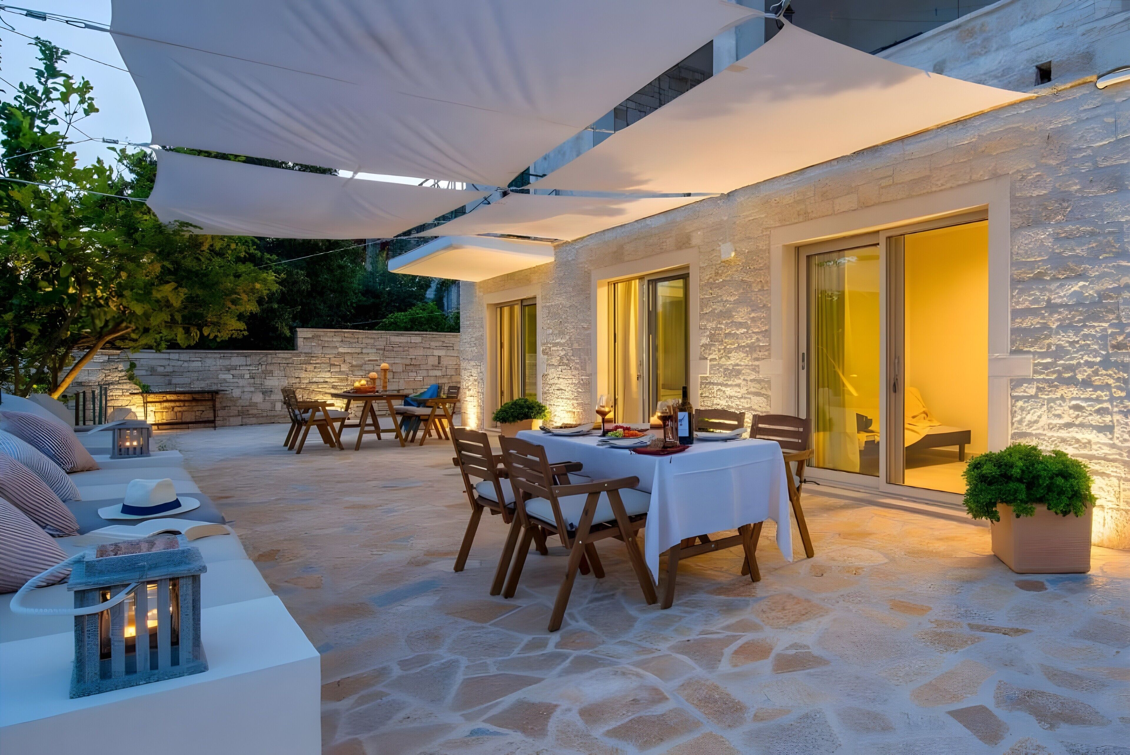 Une terrasse extérieure avec un sol en pierre, une table à manger et un coin salon agrémenté de coussins et d’un chapeau.