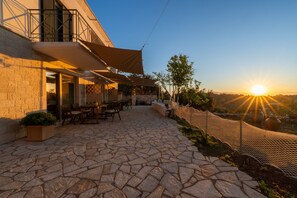 A stone-paved outdoor area with tables and chairs, a covered seating area, and a view of the sunset. - Studio Apartment 'Katerina' with Mountain View, Wi-Fi and Air Conditioning (Gaios)