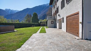 A stone pathway leading to a building with a garage, a well-maintained lawn, and mountains in the background. - Apartment 'Casa Letizia' with Private Pool, Private Terrace and Wi-Fi (Colico)