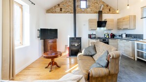 Una sala de estar con sofá, televisión, mesa de madera, pared de piedra, cocina con armarios de madera y chimenea.