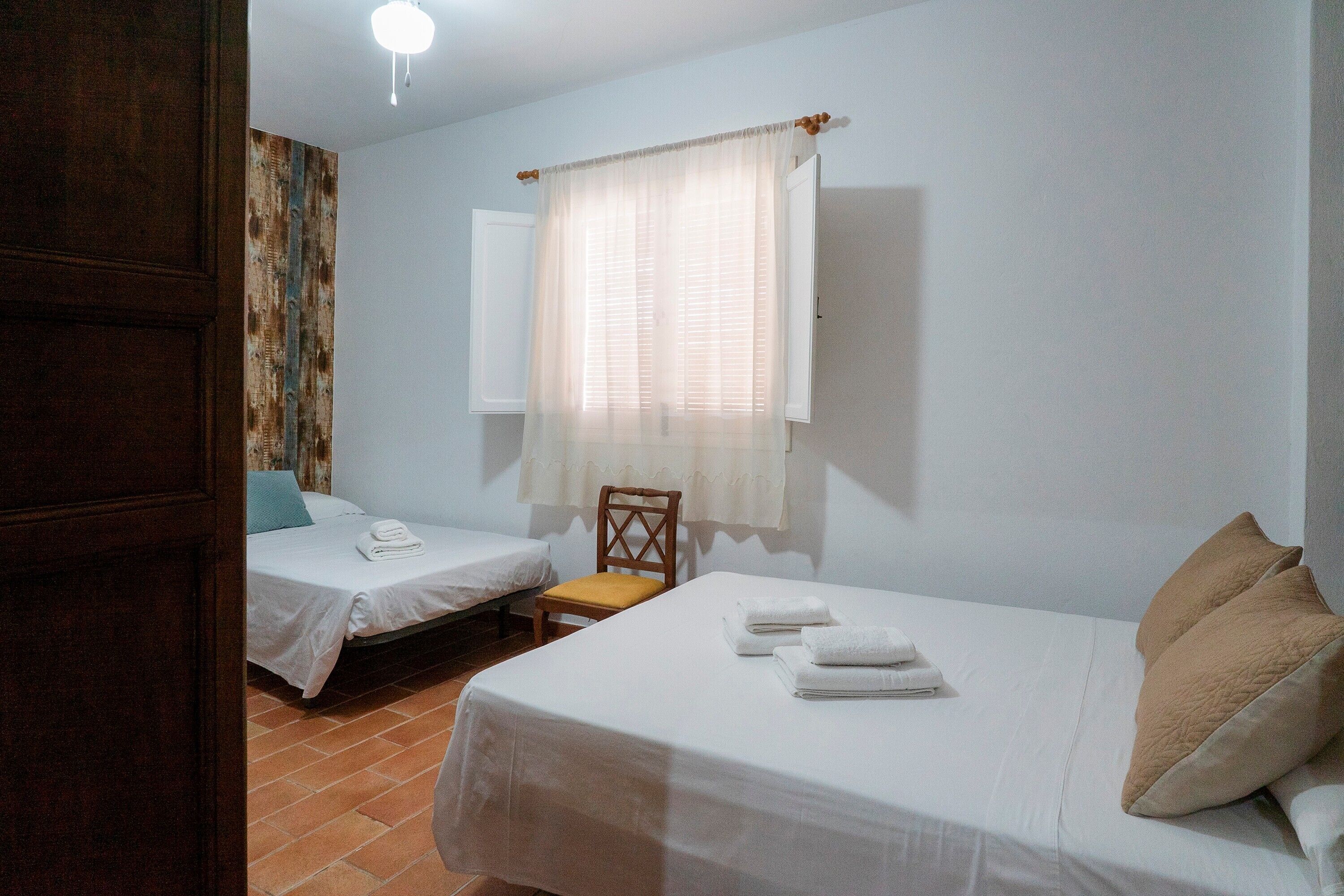 Una habitació amb dos llits, una cadira de fusta, una finestra amb cortines i una porta.