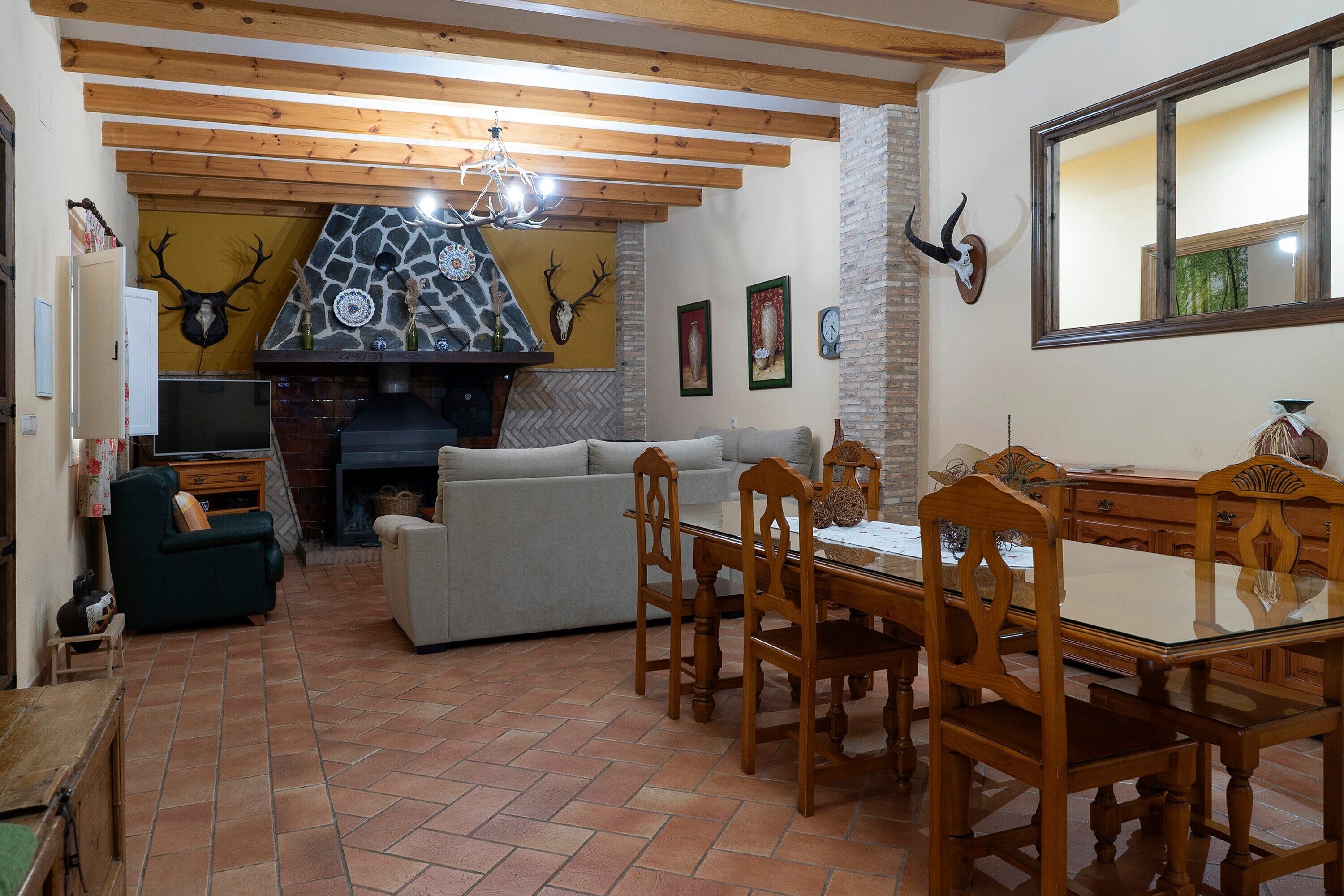 A living room with a fireplace, wooden ceiling beams, a stone wall, and a dining area with wooden chairs and a glass table.