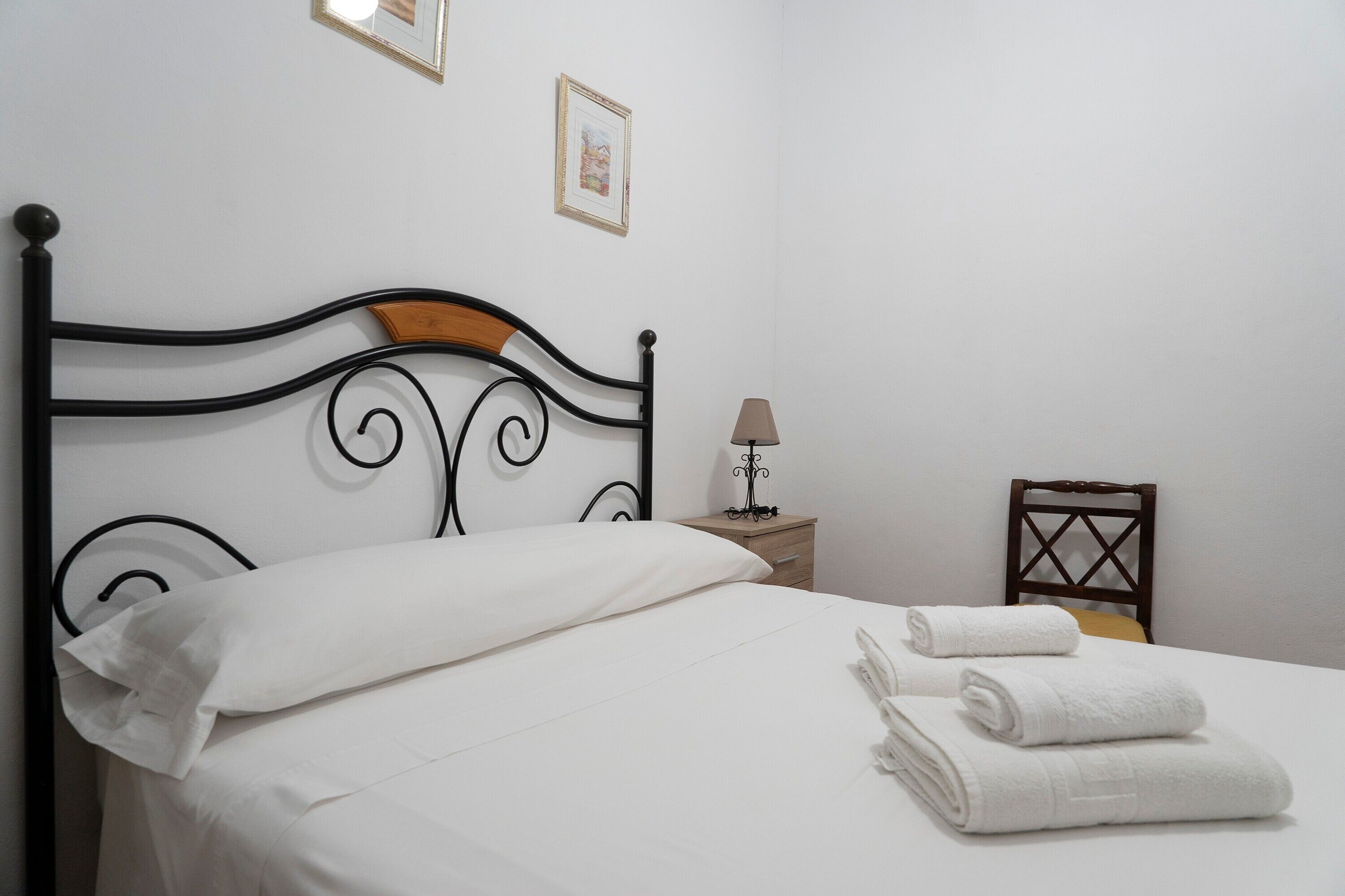 Un letto rifatto con lenzuola bianche e due asciugamani arrotolati, un comodino con una lampada, e una piccola sedia di legno.
