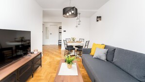 Una sala moderna con un sofá gris, una mesa de centro de madera con una planta, un televisor sobre un mueble bajo, y un comedor al fondo.