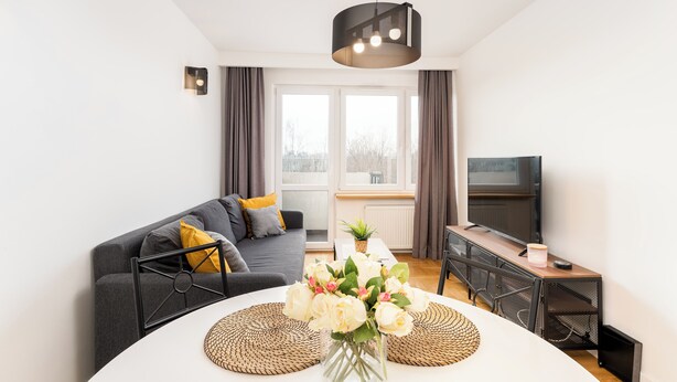 Una sala de estar moderna con un sofá gris, una mesa de centro con un jarrón de flores, un televisor y un comedor con una mesa blanca y protectores de mesa tejidos.