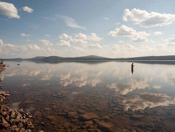 Un lago tranquilo con una persona pescando, agua cristalina que refleja el cielo y montañas lejanas.