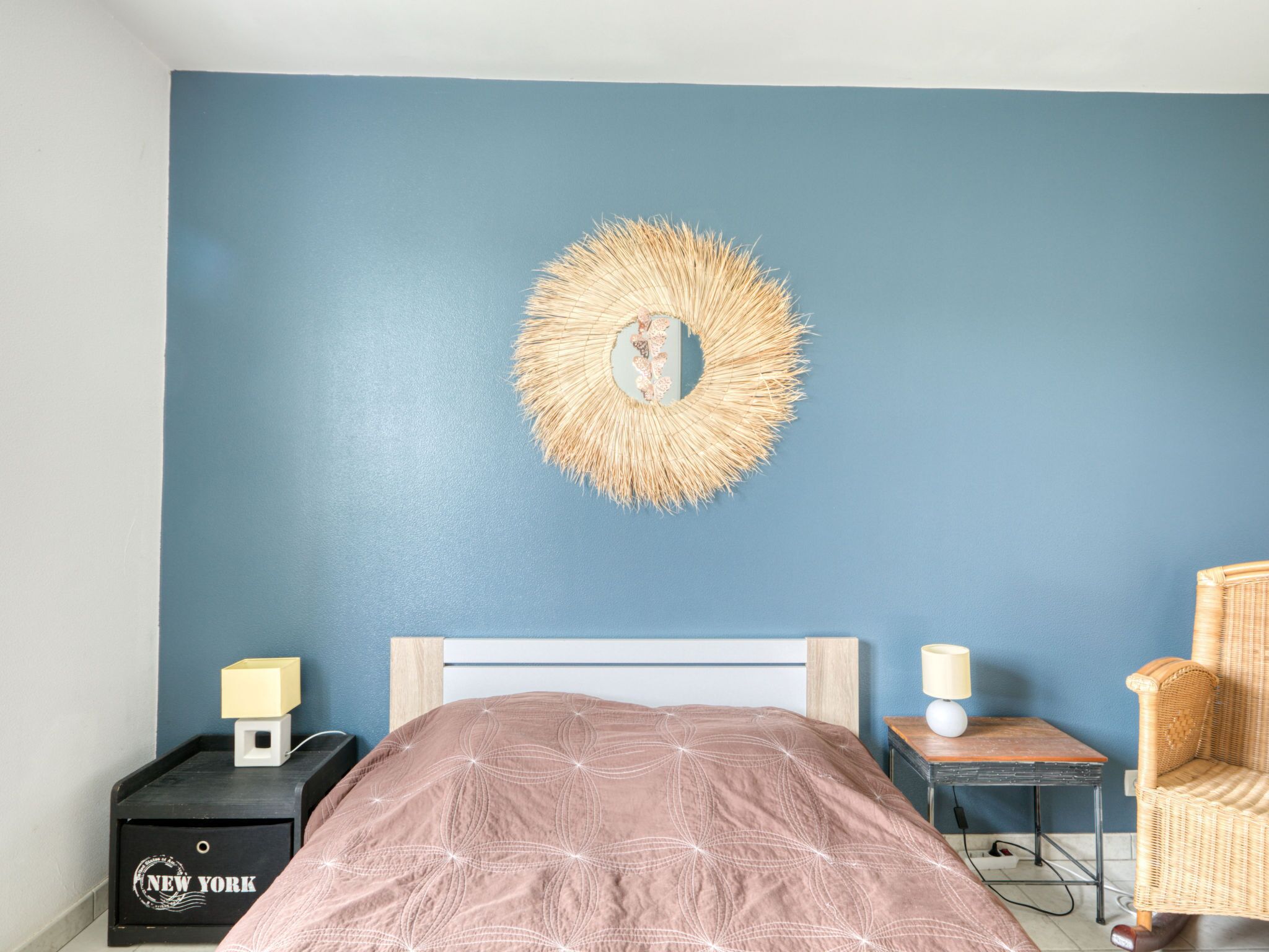 A bedroom with a blue wall, a round mirror, a bed with a patterned bedspread, a nightstand with a lamp, a wicker chair, and a small table with a lamp.