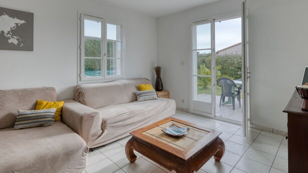 A spacious living room with a beige sofa, a wooden coffee table, and a view of a garden through large sliding glass doors. - Le Bas d'Anville by Interhome (Etaules)