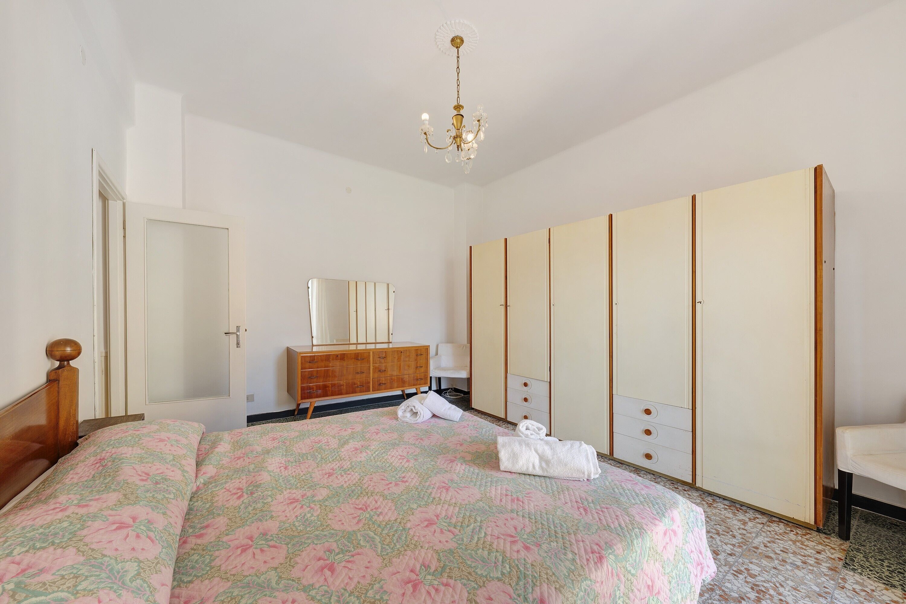 A bedroom with a bed, a wooden dresser, a wardrobe, a mirror, and a chandelier.