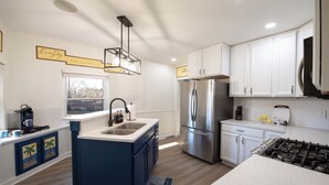 A modern kitchen with white cabinets, a stainless steel refrigerator, a gas stove, and a blue island with a sink. - The Nest:A Sanctuary of Nature & Luxury (Charlotte)