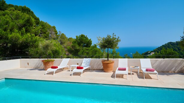 Une piscine à l’eau d’un bleu cristallin, entourée de chaises longues blanches agrémentées de coussins rouges, et offrant une vue sur la mer et les collines verdoyantes à l’arrière-plan.
