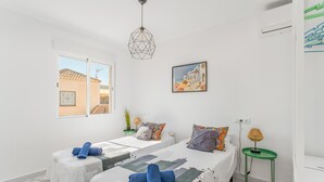 Een hotelkamer met twee bedden, een raam met uitzicht op gebouwen, een geometrisch hanglamp, een schilderij en een klein groen bijzettafeltje.