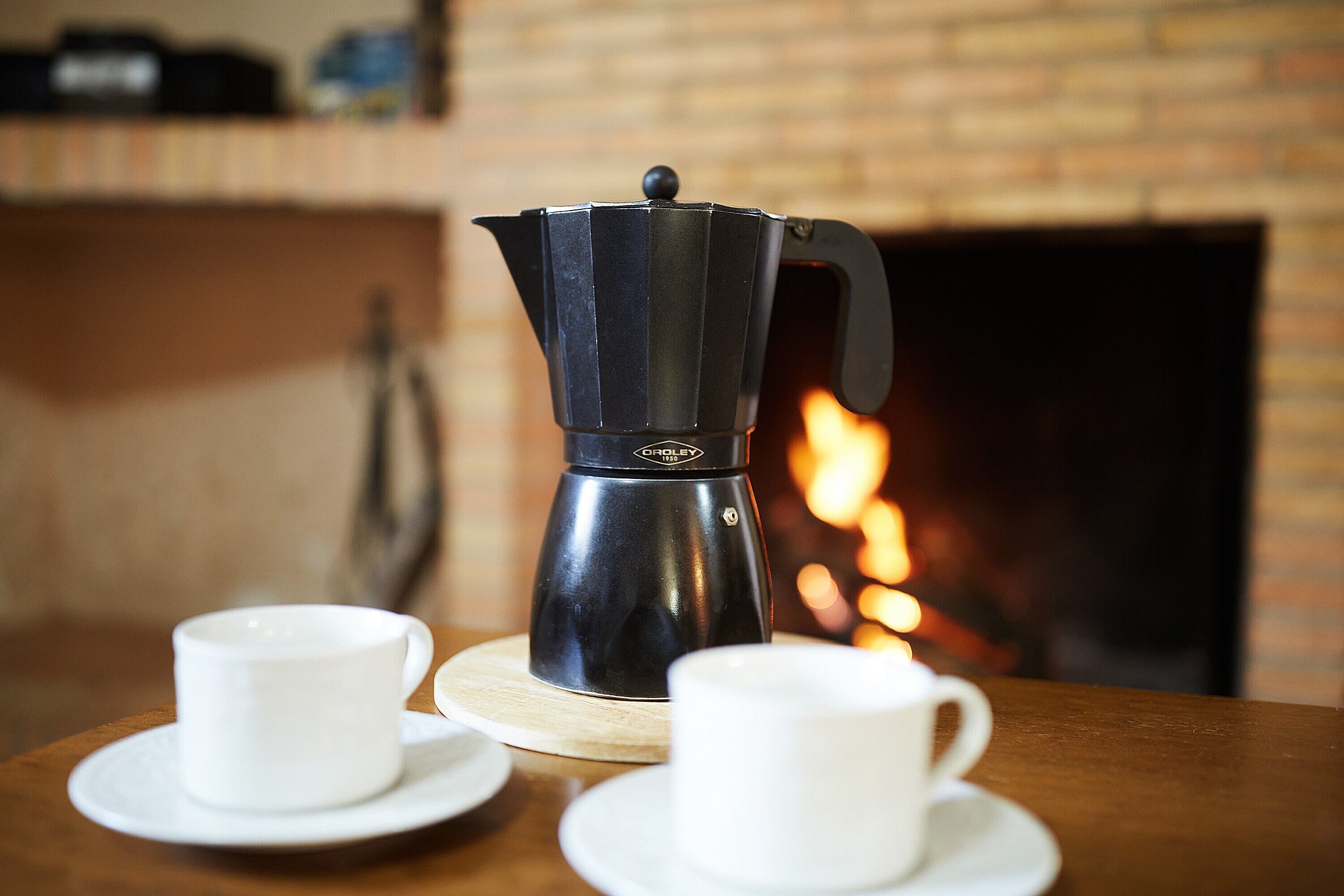 Una cafetera sobre un posavasos de madera, con dos tazas blancas sobre una mesa.