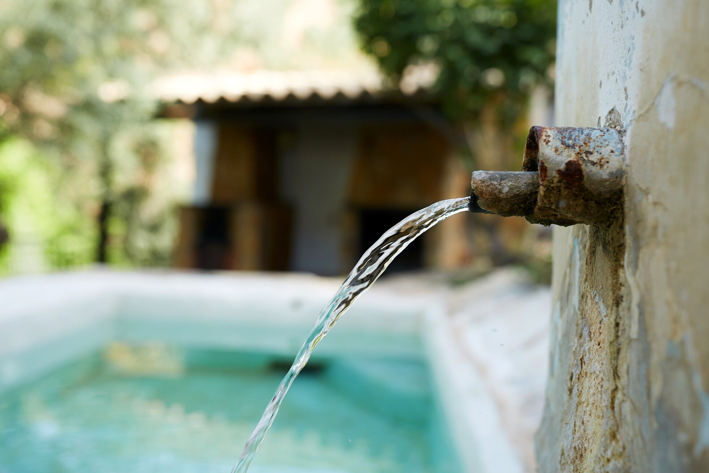 Un caño de agua con una tubería oxidada y un fondo borroso de un edificio y follaje.