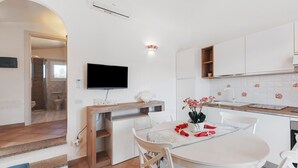 Une cuisine moderne avec des meubles blancs, une table à manger avec des chaises, un téléviseur à écran plat fixé au mur et une petite fenêtre au-dessus de l’évier.