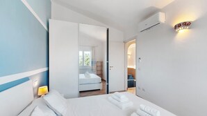 Une chambre d’hôtel avec un lit, une table de chevet, une lampe, un climatiseur et une salle de bain visible à travers une porte ouverte.