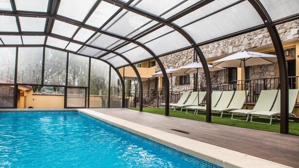 Una piscina techada con sillas de descanso y sombrillas, rodeada de muros de piedra y un techo de vidrio transparente.