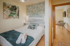 A bedroom with a bed, a green blanket, two rolled towels, a nightstand, a wall-mounted TV, and a patterned wallpaper.