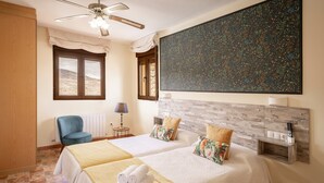 A hotel room with two beds, a ceiling fan, a large abstract painting, a blue chair, and a view of the desert through the window.